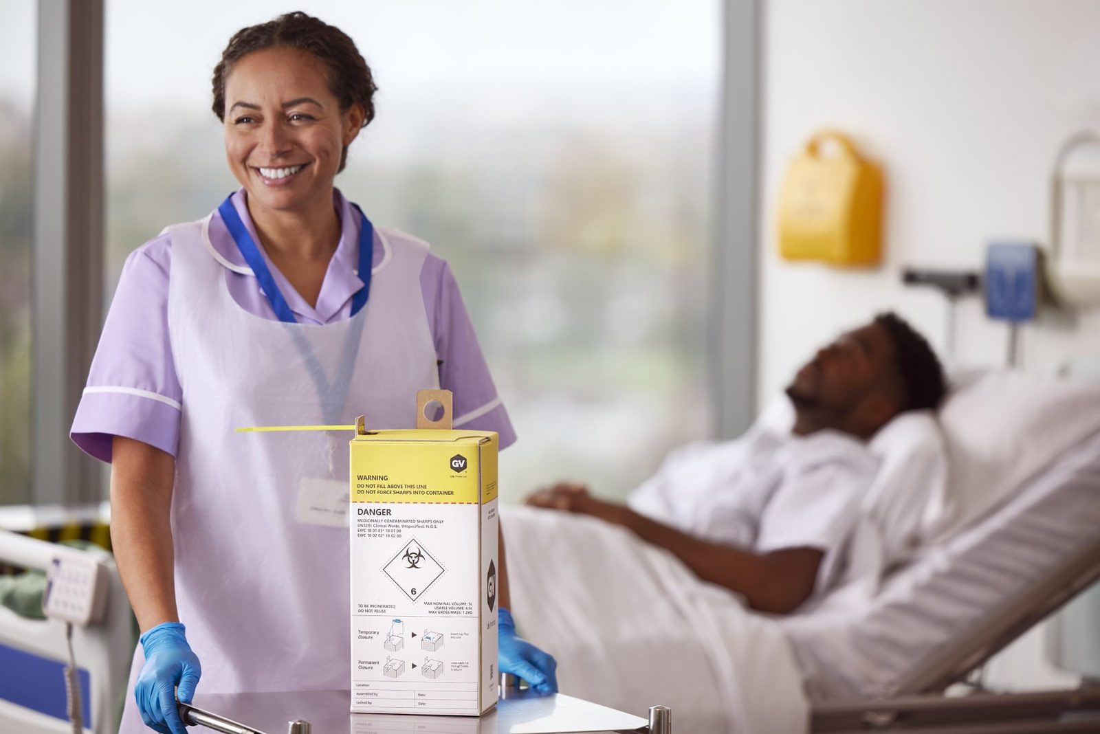 GV Health: a smiling nurse with a sharps box beside a patient in bed