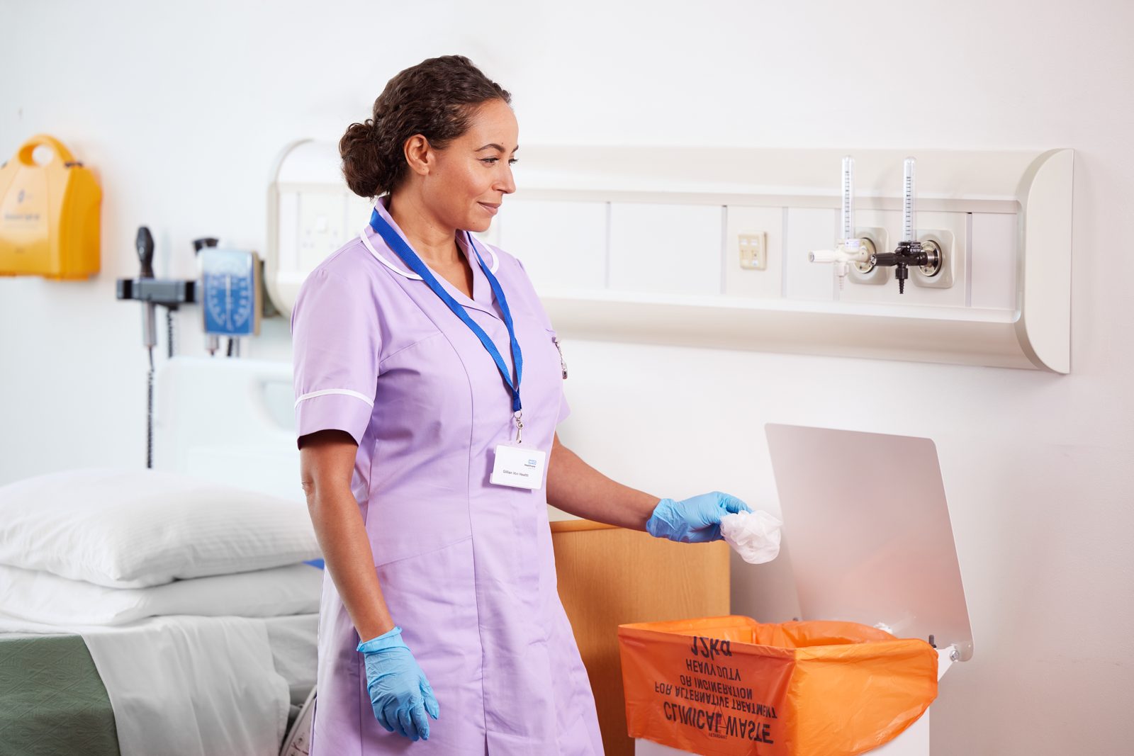 A nurse in lilac putting a used item into an orange clinical waste bag