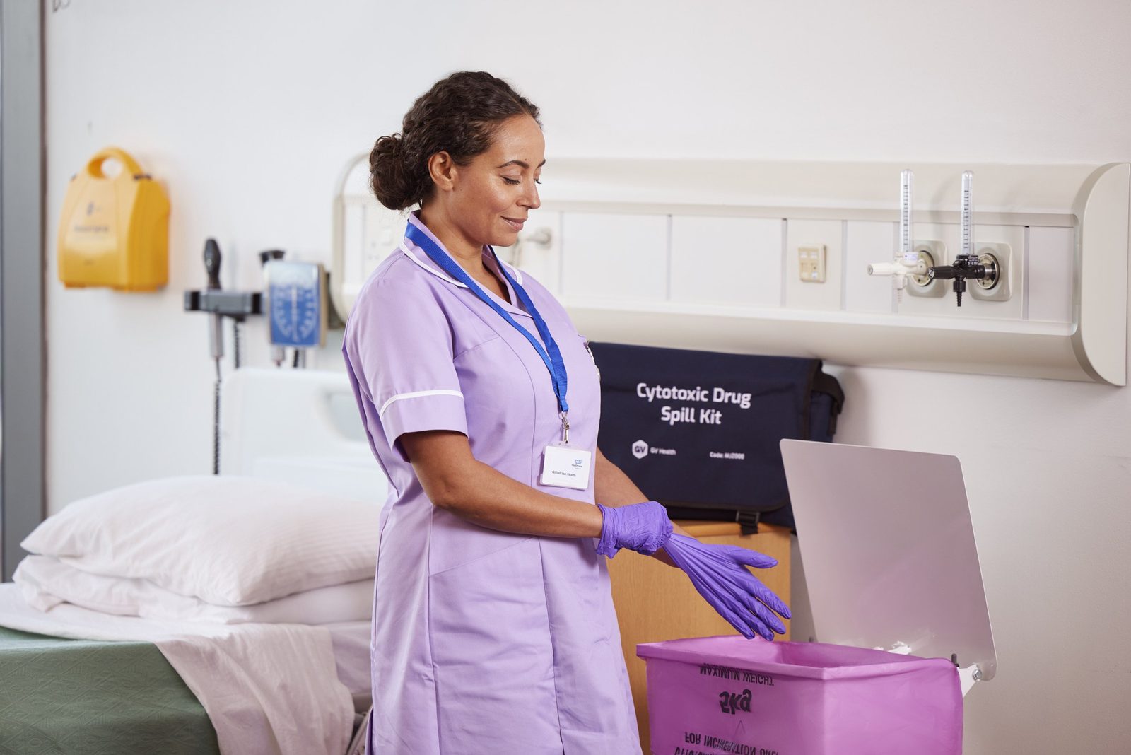 A nurse in lilac taking off purple gloves beside a clinical waste bin